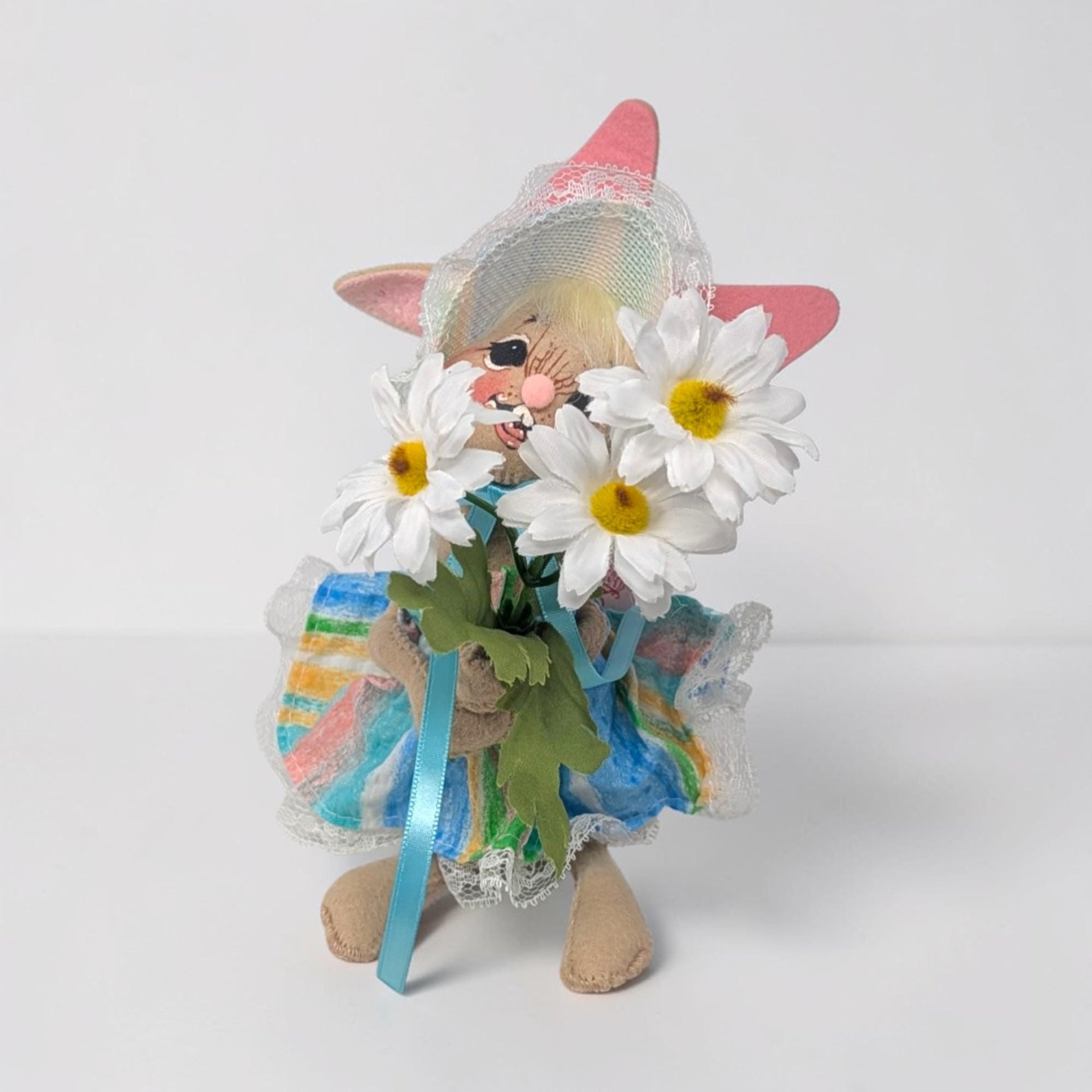 Tan felt bunny wearing a colorful striped sun hat and matching dress with white lace trim. She holds a large bouquet of three white daisies with yellow centers. Her tall ears are lined in pink felt and her face has a cheerful hand-painted smile.