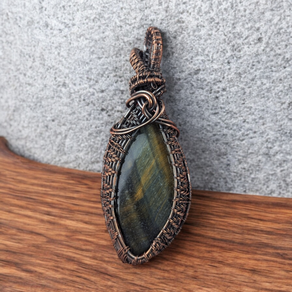 A large oval Tiger's Eye cabochon with golden-brown banded luster. It is encased in a heavy copper wire wrap with a dark oxidized finish. The wirework includes thick woven layers and multiple scrolls near a large integrated bail.
