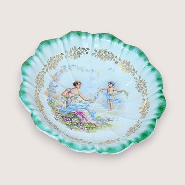 Antique Dresden porcelain bowl with a scalloped edge and green ombré rim. Center features a classical scene of a woman and a cherub with floral garlands. Gold filigree accents. 9.25 inches wide.
