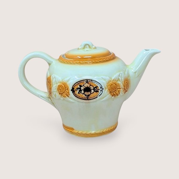 A vintage Porcelier cream-colored teapot with a raised, textured sunflower and rope border in amber-brown tones. A central black and white floral medallion sits within an oval frame. The teapot has a classic rounded shape and matching lid, set against a neutral, warm background.