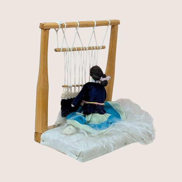 Vintage Navajo weaver doll sitting at a wooden loom. Features a miniature hand-woven textile in progress, a velvet-style bodice, and a turquoise-striped skirt on a faux-fur base. 8 inches tall.