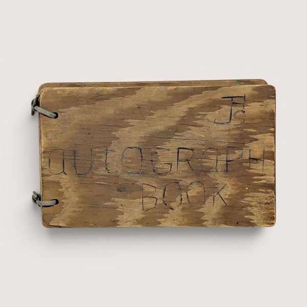 A small, rectangular autograph book with a natural wood grain cover. The words "Autograph Book" are hand-carved into the wood. Two metal rings bind the pages on the left. Rustic, weathered texture with a warm honey-oak finish.