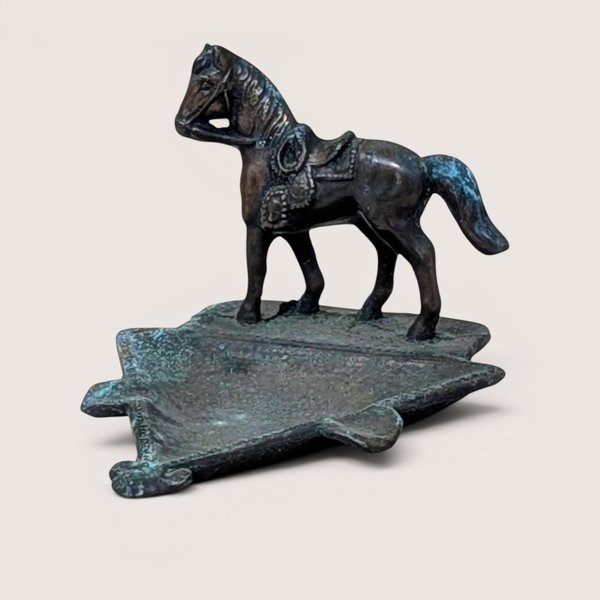 A vertical cast metal ashtray featuring a saddled horse figurine standing on a triangular base. The metal has a dark bronze patina with heavy green verdigris oxidation throughout. Sturdy, industrial texture with period-correct relief details.