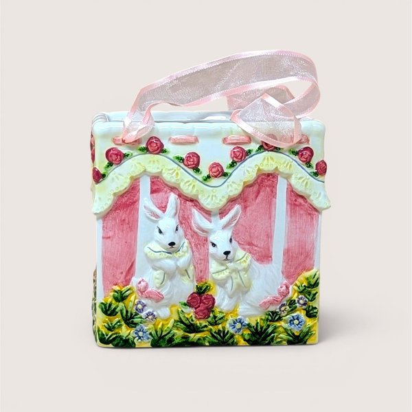 A square ceramic vase shaped like a gift bag with pink ribbon handles. The front features a high-relief scene of two white bunnies in a garden with pink roses and yellow lace detailing. Glossy vitrified texture.
