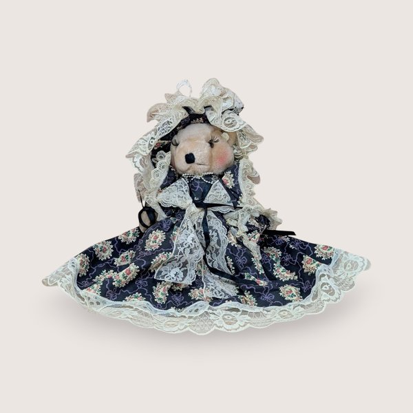 A vertical 12-inch handcrafted teddy bear with tan fur and movable limbs. Wearing an elaborate Victorian-style dark floral dress with voluminous cream lace trim and a matching oversized lace bonnet.