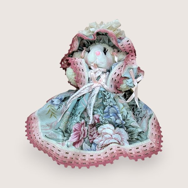 A seated plush lamb in a wide, floral-patterned sage green and rose pink dress. The lamb features long eyelashes, a pink nose, and an oversized matching lace bonnet. The intricate pink lace trim on the hem and bonnet adds a grounded, romantic texture.