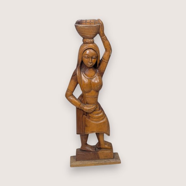 Carved wooden statue of a woman carrying a woven basket on her head, isolated on a light neutral background.