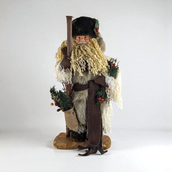 A tall, standing Father Christmas figure in a tan coat and brown fur hat. He has a long curly beard, holds a wooden staff, and carries a burlap sack with pine needles.