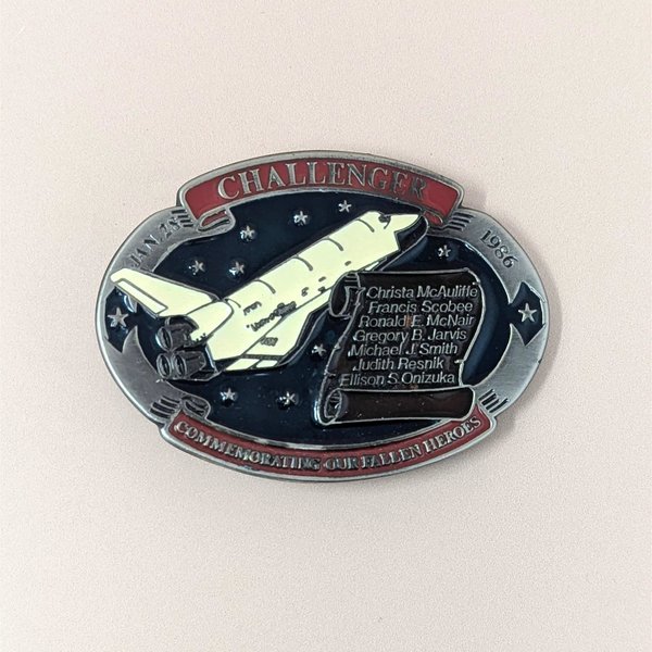 A commemorative oval belt buckle from 1986 featuring the Space Shuttle Challenger and a scroll listing the seven fallen crew members.