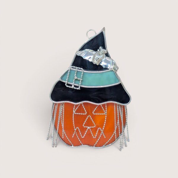 A vibrant stained glass suncatcher of a jack o' lantern wearing a black and teal witch's hat with a silver bat and wire fringe on a neutral background.