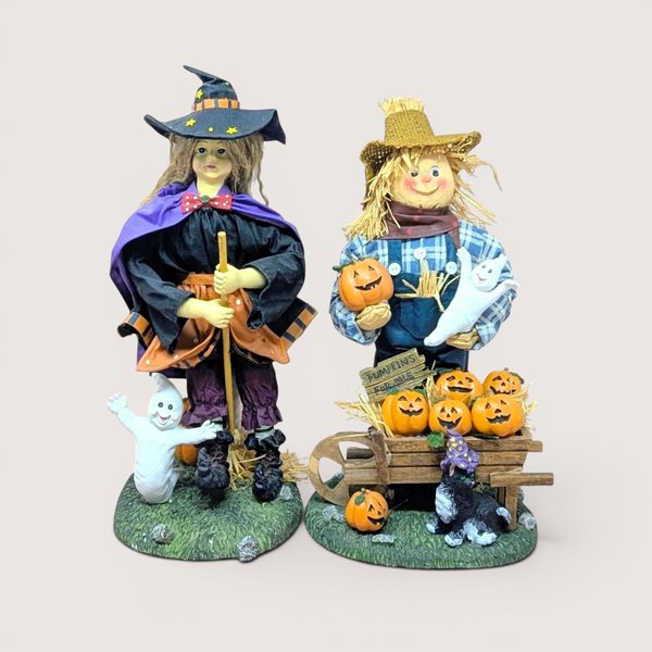 Halloween figurines of a witch with a broom and a scarecrow with a pumpkin wheelbarrow on a neutral background.