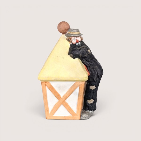 A bisque porcelain figurine of a sad clown in tattered black clothes leaning against a pale yellow street lamp against a neutral background.