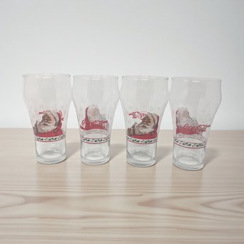 A horizontal view of four clear glass tumblers featuring the iconic Haddon Sundblom Santa Claus holding a glass of Coca-Cola. The design includes a festive holly and berry border around the base and white snowflake accents throughout the glass.