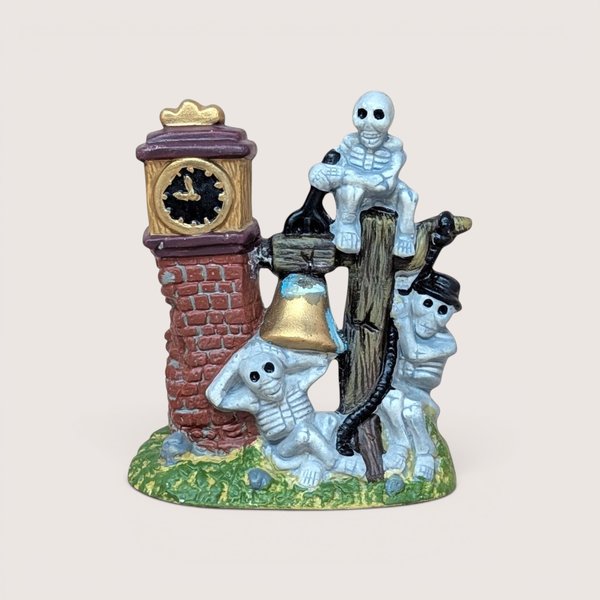 A vintage Halloween village accessory featuring a brick clock tower with three skeletons lounging around a golden bell and a wooden post.