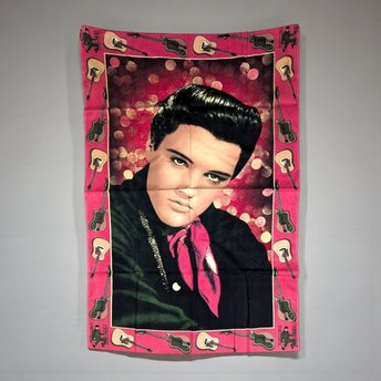 A large vertical textile wall hanging featuring a classic portrait of a young Elvis Presley wearing a pink scarf and black shirt against a shimmering red bokeh background. The piece is framed by a decorative pink border featuring alternating acoustic and electric guitars and miniature silhouettes of Elvis performing.