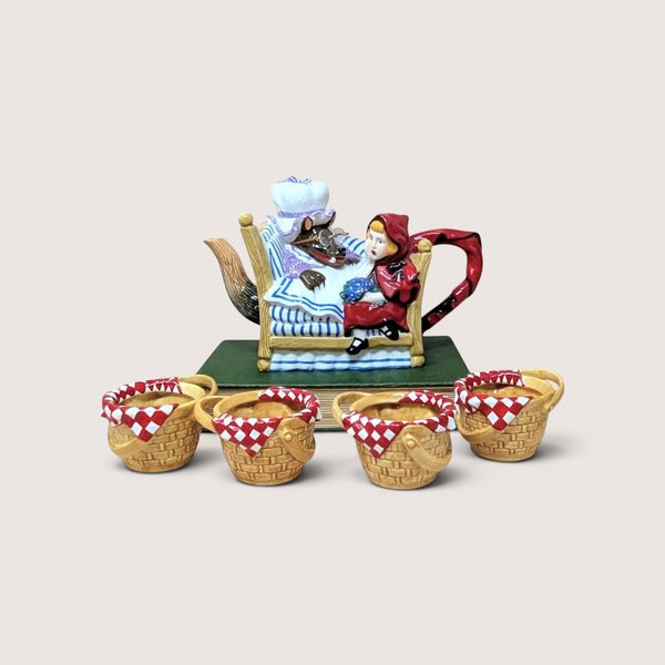 A ceramic Little Red Riding Hood teapot set with wolf in bed and four basket-shaped cups, on a beige background.