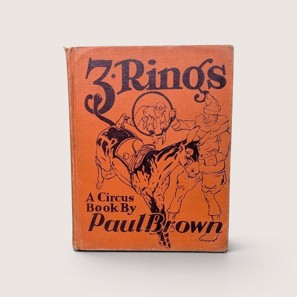 A vintage orange cloth book cover titled "3 Rings: A Circus Book By Paul Brown," centered on a beige background.
