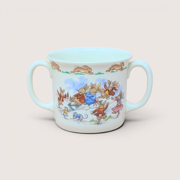 A white porcelain two-handled child's cup with illustrations of bunnies sledding, centered on a beige background.
