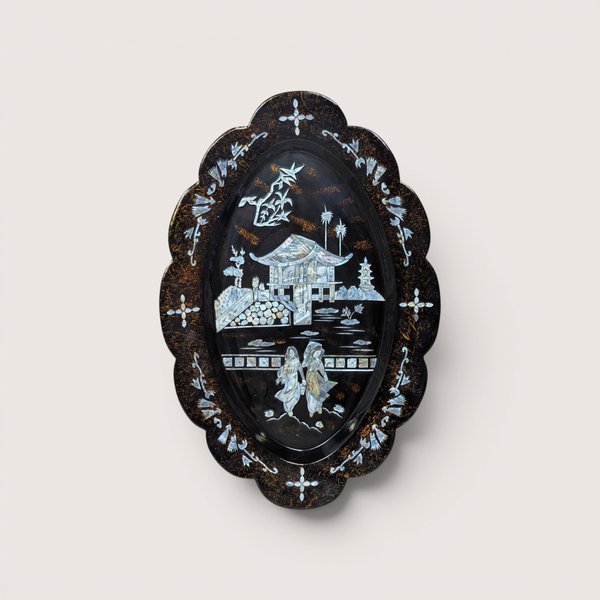 An oval black lacquer tray with mother-of-pearl inlay depicting an Asian landscape, centered on a beige background.
