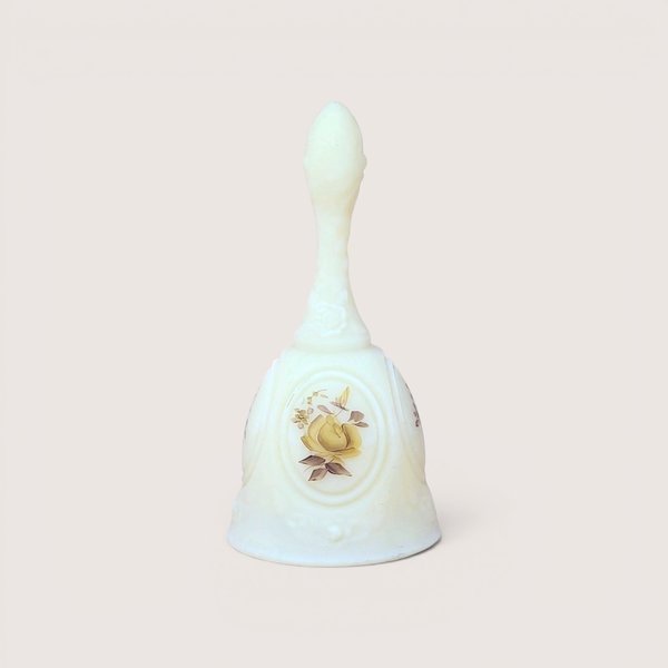 A vintage white custard glass dinner bell with a painted yellow rose design, centered on a beige background.