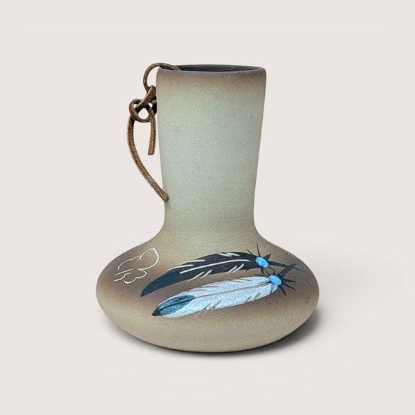 A hand painted tan pottery vase by Betty Selby featuring black and white feather motifs with turquoise accents and a leather strap tied around the neck
