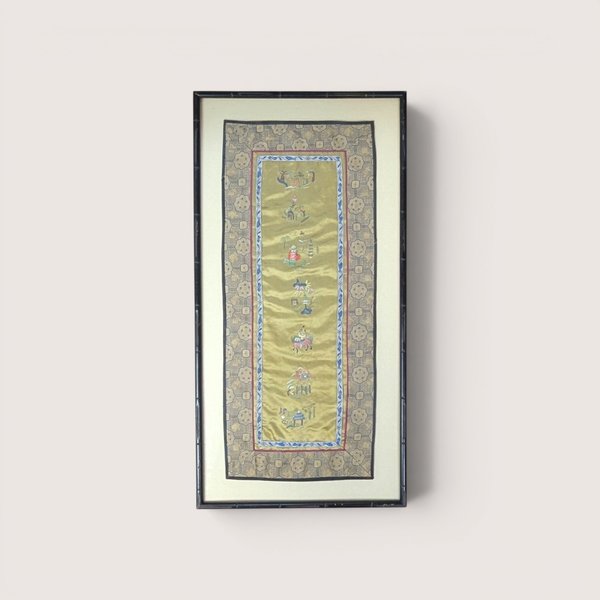 A framed vertical Chinese silk embroidery textile with small figures and decorative borders, on a beige background.