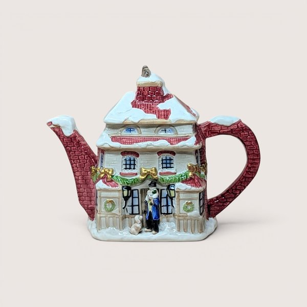 A ceramic teapot shaped like a snow-covered holiday house with festive garland, centered on a beige background.