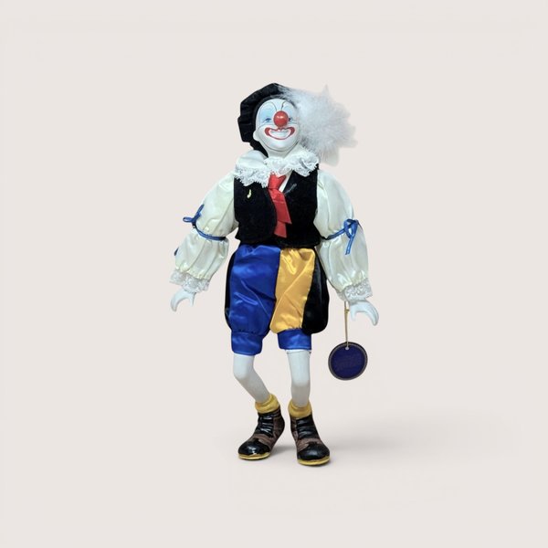 A standing clown doll with a painted ceramic face, a feathered hat, a black vest, and blue and yellow satin pants.
