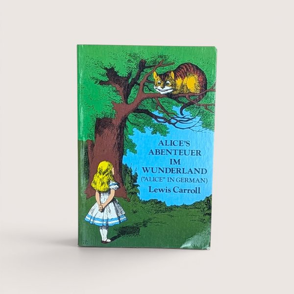 A German edition book of Alice in Wonderland featuring the Cheshire Cat in a tree, centered on a beige background.