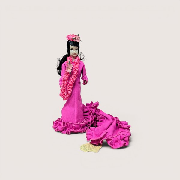 Vintage souvenir doll wearing a bright pink ruffled dress and matching pink floral lei, centered on a plain linen background.