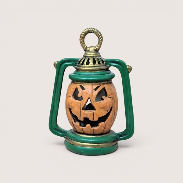 Vintage Halloween lantern decoration with a central jack-o'-lantern face, green frame, and gold trim, centered on a plain linen background.