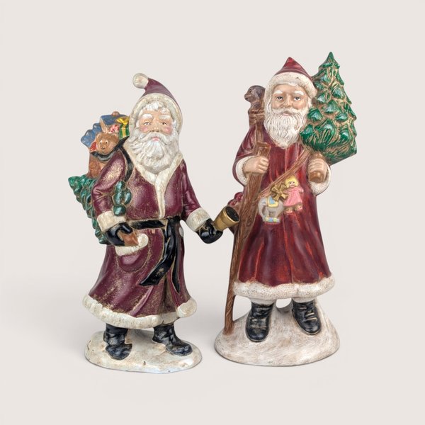 Pair of vintage hand-painted Old World Santa figurines in red coats carrying trees and toys on snowy bases.