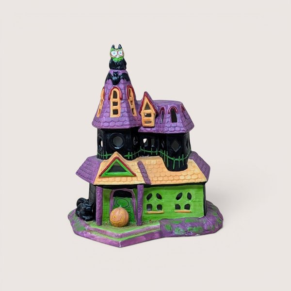 Ceramic Halloween haunted house decoration featuring purple rooftops, green accents, a jack-o'-lantern by the door, and an owl on the tower peak, centered on a plain linen background.