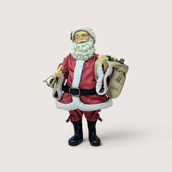 Standing Santa Claus figurine with glasses holding a gold handbell and carrying a sack of toys over his shoulder, centered on a plain linen background.