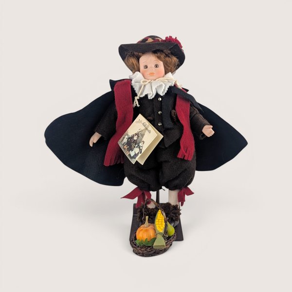 A 12.5-inch vintage Mary Michaud porcelain and fabric Samuel Eaton silhouette doll wearing a dark cape and pilgrim attire on a stand.