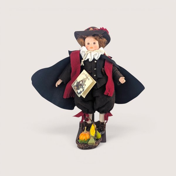 Vintage porcelain doll wearing a brown pilgrim-style outfit, black cape, and red scarf with a small basket of autumn harvest vegetables at its feet, centered on a plain linen background.