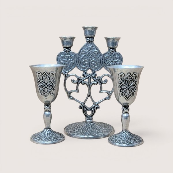 Silver-finished metal candelabra featuring intricate scrollwork and heart-shaped patterns, flanked by two matching small chalices or goblets, centered on a plain linen background.