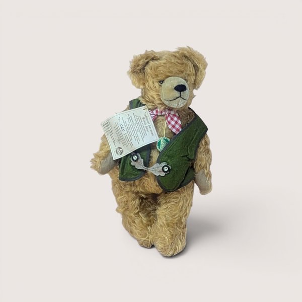Vintage mohair teddy bear wearing a traditional Bavarian green vest and red-and-white checkered bow tie, complete with its original hanging paper tag, centered on a plain linen background.