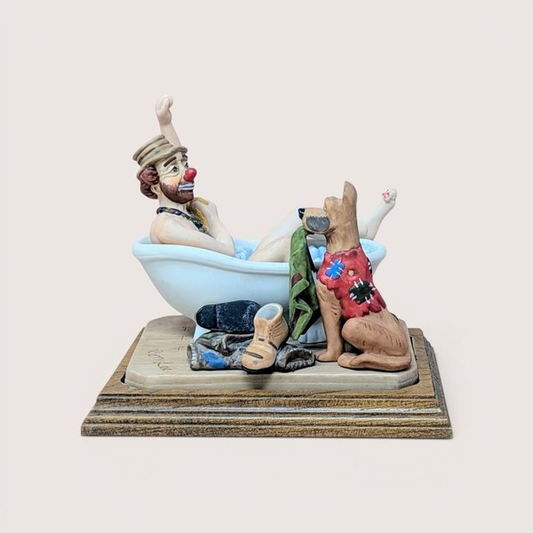 Vintage Ron Lee Emmett Kelly Jr. hobo clown figurine depicting the clown in a white clawfoot bathtub with a dog sitting alongside holding his green towel, mounted on a wooden base and centered on a plain linen background.