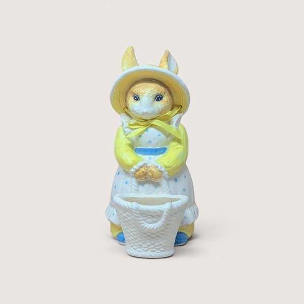 Vintage ceramic bunny figurine dressed in a yellow long-sleeved shirt, a yellow bonnet with a bow, and a blue polka-dot apron dress, standing behind a small woven white basket, centered on a plain linen background.