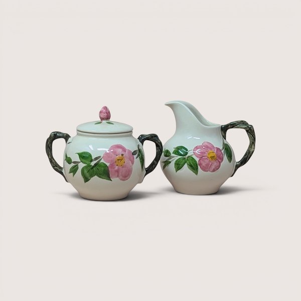 Vintage ceramic sugar bowl and creamer set featuring hand-painted pink wild roses and green leaves on a cream background, complete with twig-style handles and a rosebud finial on the sugar bowl lid, centered on a plain linen background.