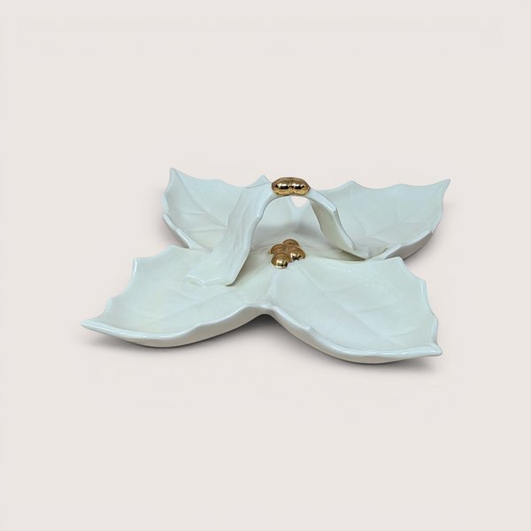 Vintage white ceramic divided serving dish shaped like a four-lobed holly leaf cluster, featuring a central looped leaf handle accented with gold-gilt berries at the top and base, centered on a plain linen background.