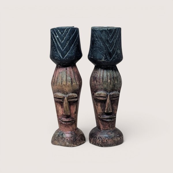 Pair of vintage hand-carved wooden candlesticks depicting stylized elongated faces with carved hair details, each supporting a dark-stained cup-style candle holder accented with geometric chevron patterns, centered on a plain linen background.