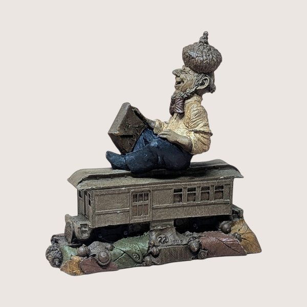 Vintage Tom Clark Cairn studio gnome figurine depicting a gnome named "Cole" wearing an acorn-cap hat, seated on top of a miniature trolley car or train passenger car numbered 22, resting on a base of textured leaves and acorns, centered on a plain linen background.