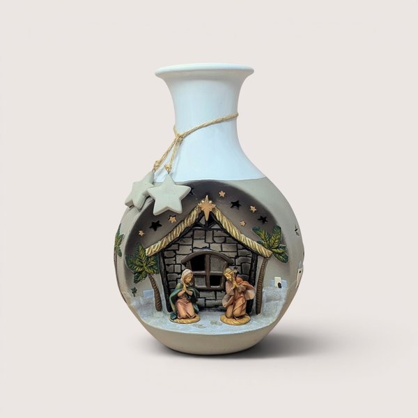 Vintage ceramic vase featuring a built-in, recessed Nativity scene on the front, depicting Mary and Joseph kneeling inside a stone stable flanked by palm trees under cutout star accents, complete with a white glazed upper neck wrapped in natural twine with hanging clay stars, centered on a plain linen background.