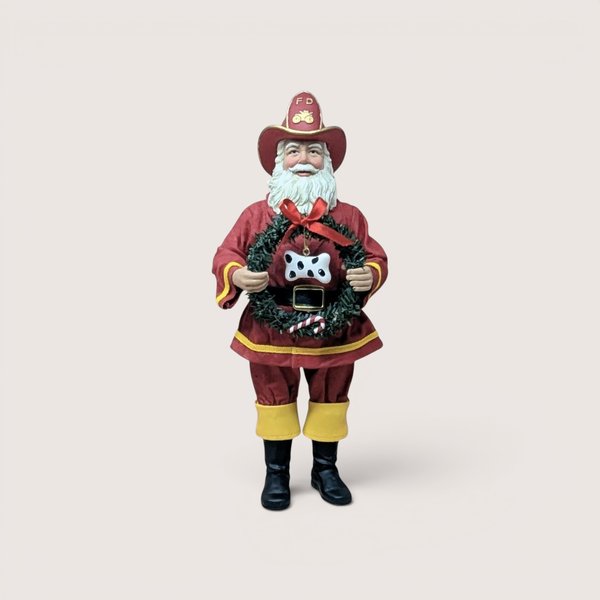 Vintage standing Santa Claus figurine dressed as a firefighter in a red turnout coat with yellow trim, black boots, and a matching red "FD" firefighter hat, holding a green Christmas wreath adorned with a red bow, a candy cane, and a black-and-white spotted dog bone ornament, centered on a plain linen background.