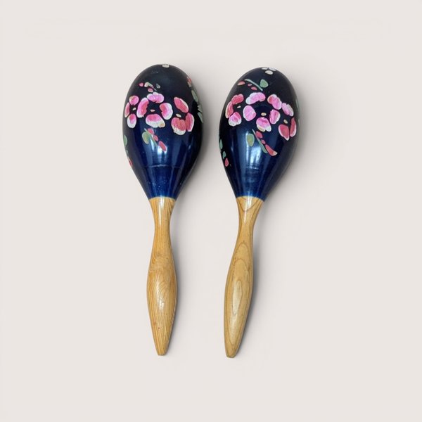 A pair of vintage hand painted wooden maracas on a soft pink background. The musical instruments feature sleek natural wood grain handles and glossy dark blue bulbs decorated with hand painted bright pink and red flowers.