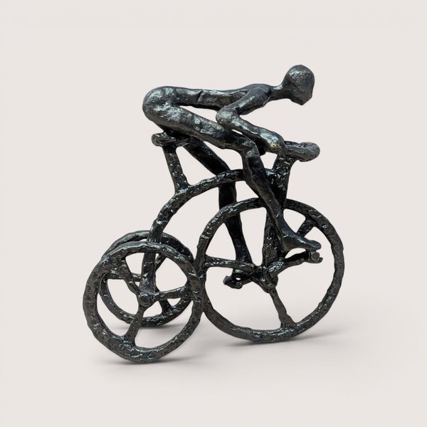 A vintage brutalist cast iron sculpture of a man riding a tricycle on a soft pink background. The dark, heavily textured metal figurine depicts a stylized, faceless human figure leaning forward in a racing position over three spoked wheels.
