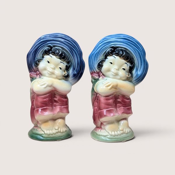 A pair of vintage Royal Copley ceramic planters on a soft pink background. Made in the 1950s, each matching vase features a charming barefoot girl wearing a massive blue sunbonnet and a pink dress with a glossy, airbrushed finish.