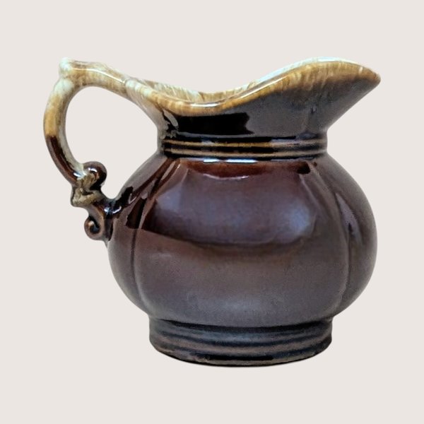A vintage small ceramic pitcher with a bulbous body and a glossy brown drip glaze handle and rim on a plain background